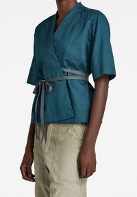 Personne à la peau foncée portant un haut cache-cœur bleu sarcelle avec une ceinture grise et un pantalon beige clair, debout devant un fond blanc uni.