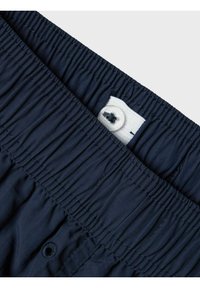 Los pantalones de baño en color navy cuentan con una cinturilla elástica con textura recogida, un botón blanco y una pequeña etiqueta cosida en el interior.