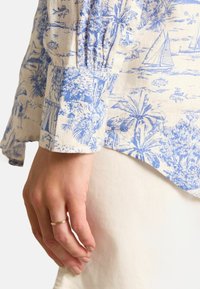 Lewa ręka z cienką złotą obrączką, nosząca niebieską i białą koszulę z długim rękawem oraz białe spodnie na jednolitym tle.