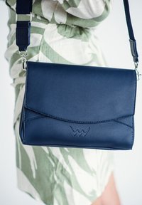 Modrá kožená crossbody taška s překlápěcí klopou, zdobená dekorativním logem, stříbrnými detaily a nastavitelným popruhem.