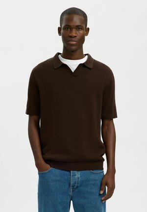 Giovane uomo con capelli corti che indossa una polo marrone scuro a maniche corte sopra una maglietta bianca e jeans blu, in piedi davanti a uno sfondo semplice.