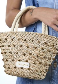 Bolsa de paja tejida con acentos de cuentas doradas, forma ovalada, dos asas trenzadas y una etiqueta de logo de Gioseppo en la parte frontal.