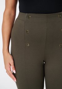 Main de femme près d'un pantalon taille haute vert olive avec quatre boutons dorés visibles et un haut noir rentré.