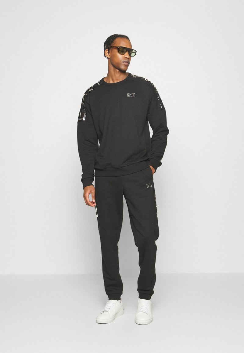 Sudadera de algodón negra y joggers, con un logo en el pecho y detalles estampados en los hombros, combinados con zapatillas blancas.