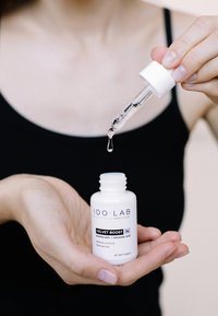 Biała cylindryczna butelka z etykietą "IDO LAB" i napisem "VELVET BOOST". Kroplomierz z klarownym serum, trzymany w dłoni, na neutralnym tle.