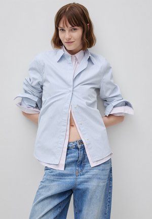 Mujer joven con pelo corto y castaño que lleva una camisa de rayas azul claro sobre una camiseta rosa y unos vaqueros azules holgados, de pie frente a una pared gris lisa.
