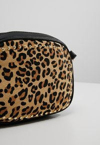 Leopardikuvioinen pussukka, jossa mustat reunat ja pienet pyöreät kultaiset niitit edessä olevan paneelin ympärillä, lepää vaaleanharmaalla pinnalla.