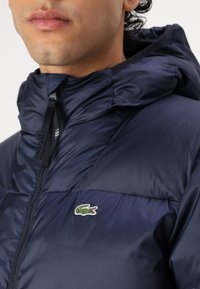 Námornícka bunda s kapucňou typu puffer so zipsom, s malým zeleno-bielym vyšitým logom na ľavej hrudi a textúrovanou tkaninou.