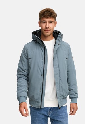 Ung mand iført en lyseblå polstret jakke med hætte, hvid skjorte og blå jeans, som står foran en ensfarvet lys baggrund.