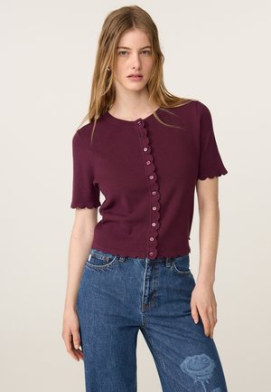 Jonge vrouw met lang haar die een bordeauxrode, korte mouwen en knopen heeft en een blauwe spijkerbroek met een rozenontwerp op de dij draagt.