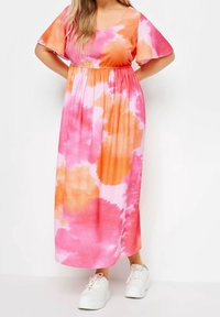 Maxi šaty v růžovém a oranžovém tie-dye, s krátkými křidélkovými rukávy a elastickým pasem, doplněné bílými teniskami.