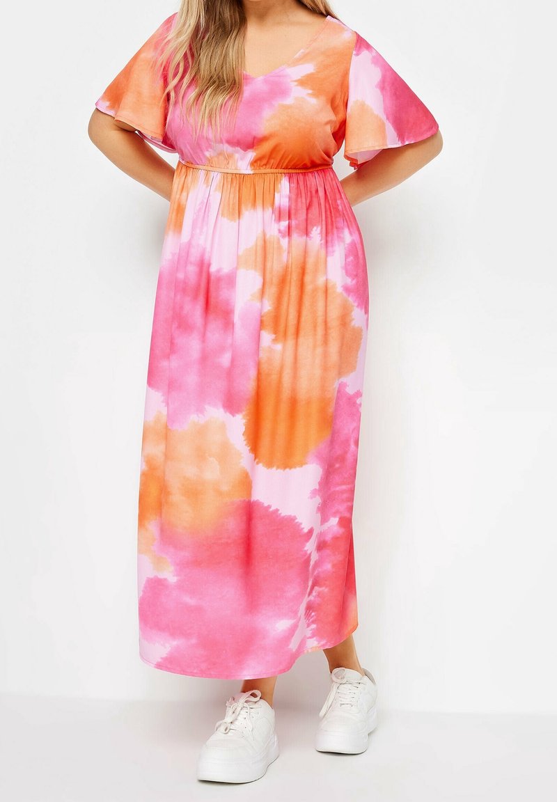 Maxi šaty v růžovém a oranžovém tie-dye, s krátkými křidélkovými rukávy a elastickým pasem, doplněné bílými teniskami.