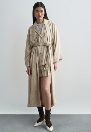 Robe longa beige com padrão e atilho, sobre uma blusa cinza e calções a condizer. Meias de lã castanhas e saltos brancos pontiagudos completam o visual.