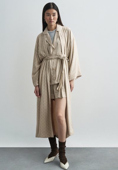 Túnica larga de color beige con un patrón y un lazo, superpuesta a una blusa gris y pantalones cortos a juego. Medias de punto marrones y zapatos de tacón blanco en punta completan el conjunto.