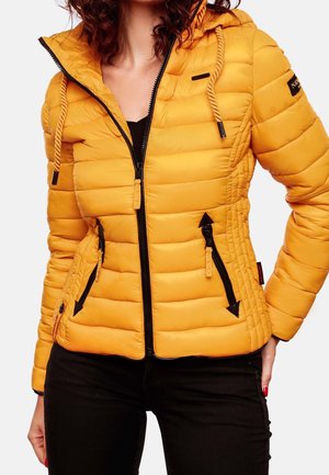 Kobieta nosząca dopasowaną żółtą pikowaną kurtkę z czarnym zamkiem błyskawicznym, kapturem i czarnymi spodniami, stoi z jedną ręką na biodrze.
