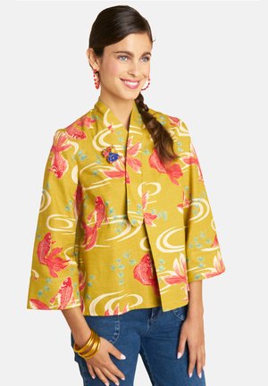 Woman with dark braided hair wears yellow kimono-style top with red fish pattern, blue jeans, red earrings, and gold bangles, smiling slightly.