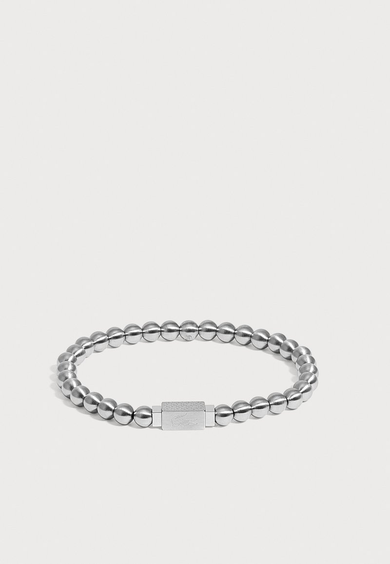 Bracelet en argent avec des perles rondes en métal et un fermoir rectangulaire gravé d'un logo de crocodile sur un fond uni.