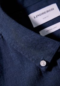 Camicia in cotone-lino navy con vestibilità slim, tessuto testurizzato, colletto a punta e un unico bottone al collo. Etichetta del brand visibile.