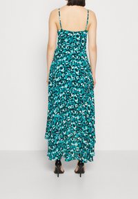 Robe maxi en turquoise avec un motif de formes abstraites. Dotée de fines bretelles et d'un ourlet fluide. Associée à des chaussures à talons hauts noires.