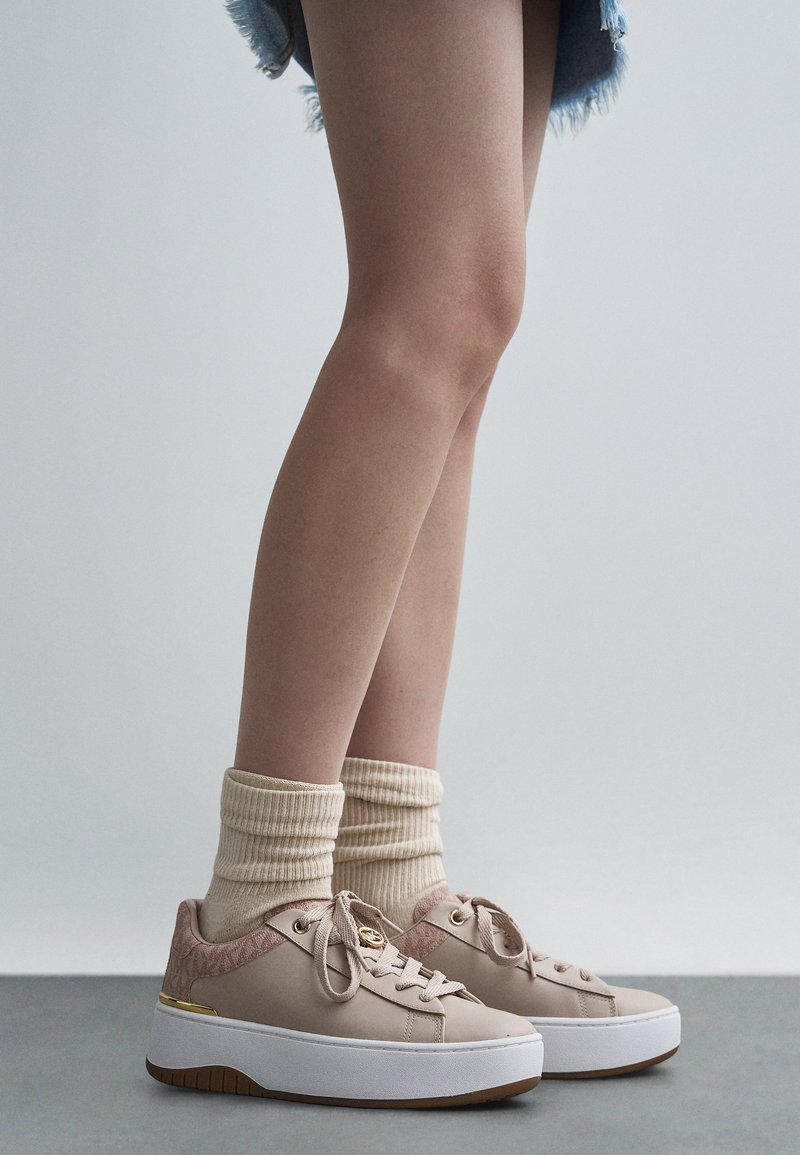 Világos rózsaszín platform sportcipők texturált díszítésekkel, arany színű fém részekkel és fűzőkkel, krémszínű bordázott zoknival és farmer rövidnadrággal párosítva.