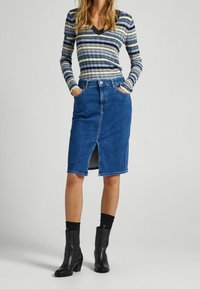 Jeans-Bleistiftrock in Mittelblau mit einem frontalen Schlitz, Fünf-Taschen-Design, Knopfverschluss, kombiniert mit einem gestreiften Langarmoberteil und schwarzen Stiefeletten.