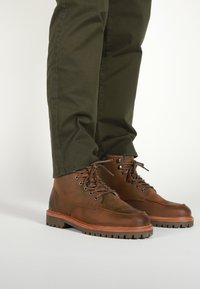 Stivali di pelle marrone con suole robuste, design con lacci, punte arrotondate e dettagli testurizzati sulla parte superiore. Abbinati a pantaloni verde oliva.