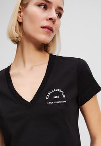 Juodas medvilninis marškinėlis su V formos iškirpte, kairėje pusėje siuvinėtas baltas tekstas: "KARL LAGERFELD PARIS 21 RUE ST-GUILLAUME".