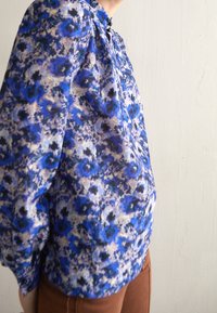 Blouse fleurie bleue avec un col montant, tissu froncé et manches longues. Le motif présente des nuances de bleu et de noir sur un fond clair.