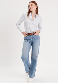 Jeans droits bleu clair associés à une chemise blanche à boutons avec de subtiles rayures bleues, complétés par une ceinture marron et des baskets blanches.