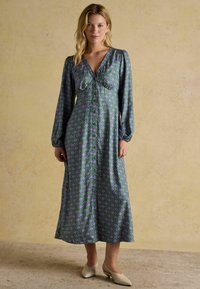 Dlhé šaty s gombíkmi v purpurovej a zelenej geometrickej vzore. Má hlboký výstrih do V, dlhé rukávy a splývavý strih.