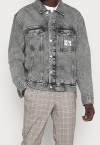 En person bär en grå denimjacka med en logotyp och rutiga byxor, stående mot en vit bakgrund.