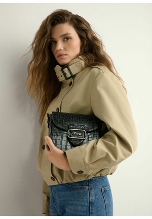 Kobieta z długimi falowanymi włosami, ubrana w beżową kurtkę i niebieskie dżinsy, trzymająca czarną torebkę z fakturą i srebrną klamrą.