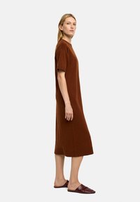 Brązowa sukienka o krótkim rękawie, długości do kolan, w luźnym kroju, wykonana z miękkiego materiału. Modelka nosi bordowe buty wsuwane. Gładka tekstura i prosty design.