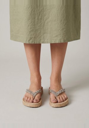 Füße in beigen, geflochtenen Plateauflipflops mit Strassriemen, gezeigt unter einem knielangen olivgrünen Rock auf neutralem Hintergrund.