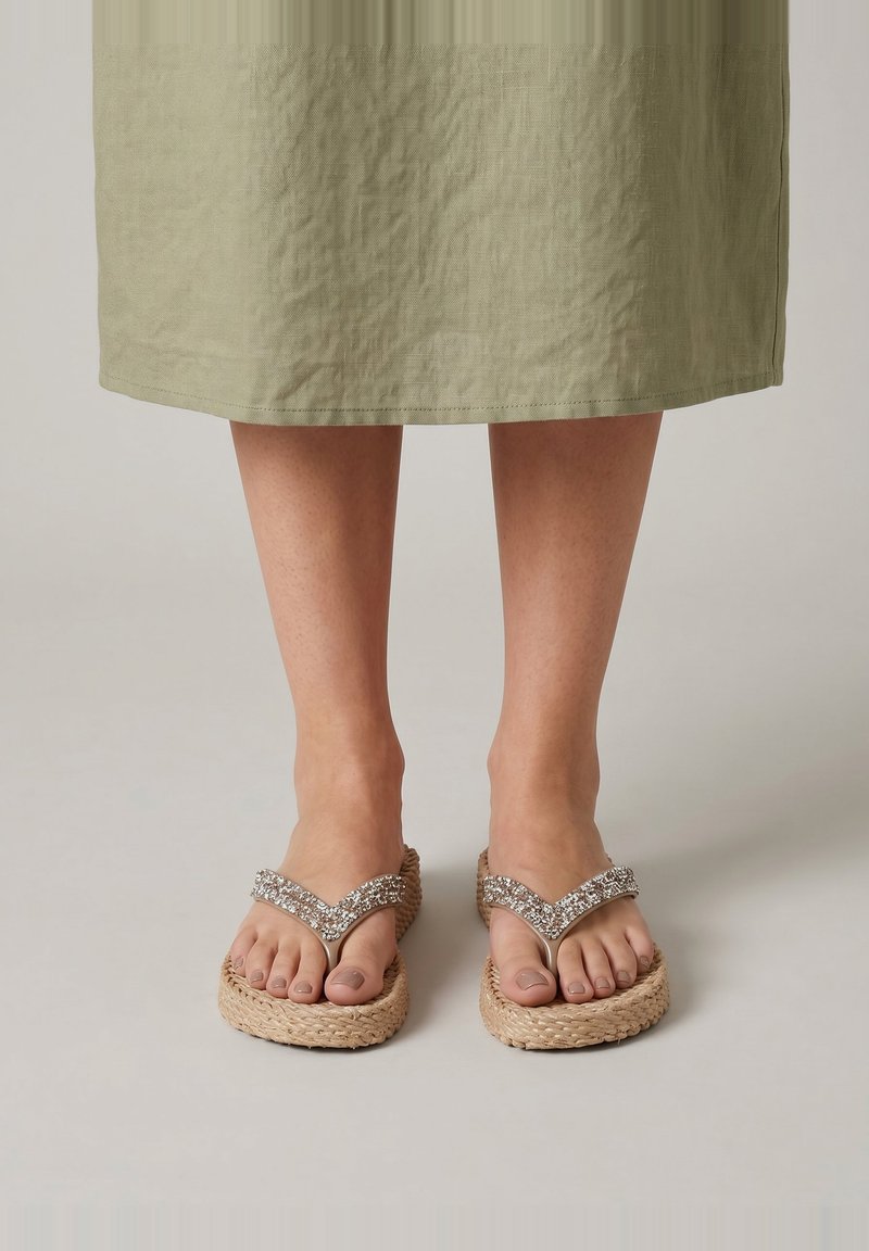 Füße in beigen, geflochtenen Plateauflipflops mit Strassriemen, gezeigt unter einem knielangen olivgrünen Rock auf neutralem Hintergrund.