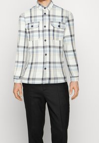 Camicia di flanella a scacchi in azzurro chiaro, grigio e bianco con due tasche sul petto e bottoni neri. Maniche lunghe e design del colletto classico.