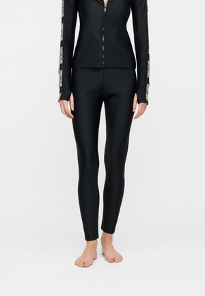 Femme portant des leggings noirs moulants et un top à manches longues zippé assorti avec le texte blanc « MOSCHINO » sur les manches, pieds nus.