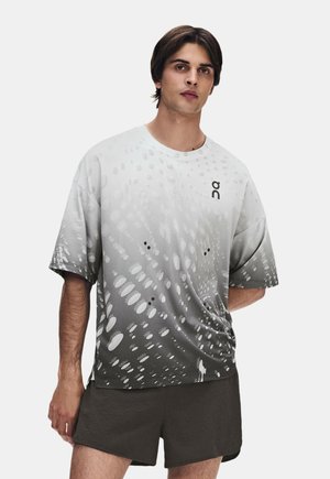 Junger Mann, der ein weißes und graues T-Shirt mit Punkte-Muster und dunkle Shorts trägt, steht vor einem schlichten hellen Hintergrund.
