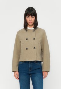 Kvinne iført en beige dobbeltspent kort jakke over hvit skjorte og blå jeans, stående mot en ensfarget hvit bakgrunn.