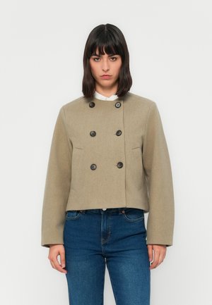 Kvinne iført en beige dobbeltspent kort jakke over hvit skjorte og blå jeans, stående mot en ensfarget hvit bakgrunn.
