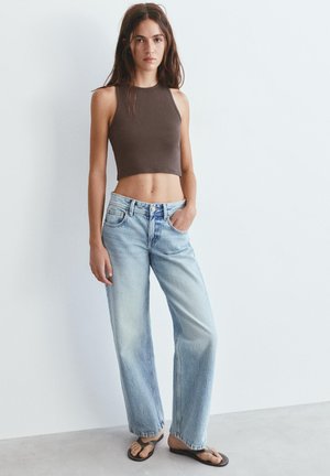 Jeune femme debout avec une main dans la poche portant un top court marron sans manches, un jean large bleu clair et des tongs noires.