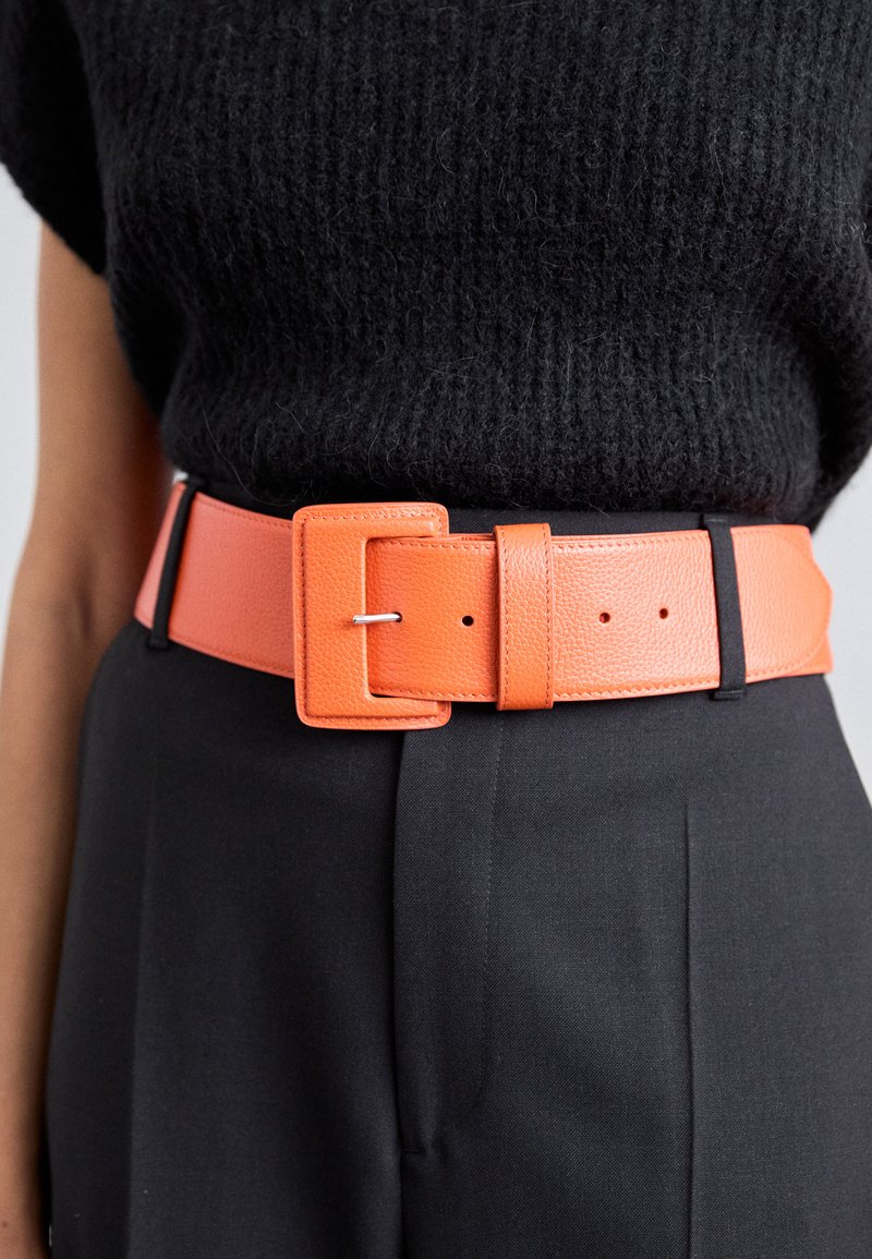 Cintura in pelle arancione con fibbia rettangolare e finitura testurizzata, indossata su pantaloni neri, caratterizzata da fori equidistanti per la regolazione.