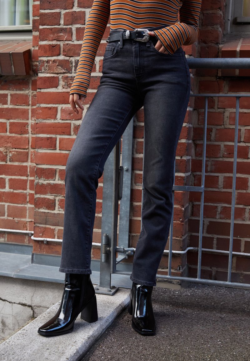 Jeans in denim scuro a vita alta con una vestibilità slim, abbinati a stivali neri lucidi alla caviglia con punta quadrata e tacco a blocco.