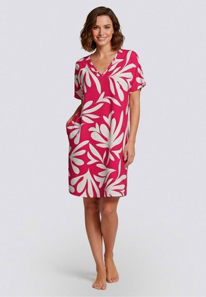 Nainen seisoo paljain jaloin yllä punainen polvipituinen mekko, jossa on suuret valkoiset lehtikuviot ja lyhyet hihat, hymyillen ja käsi taskussa.