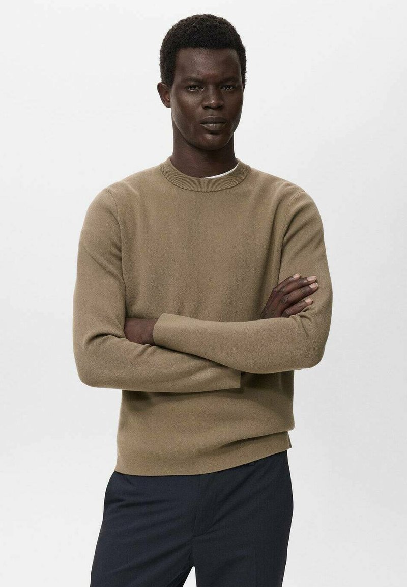 Pull en tricot beige avec un col rond et des manches longues. La texture est lisse et ajustée, associé à un pantalon bleu marine.