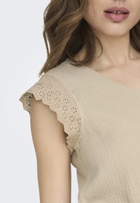 Haut beige avec des manches en dentelle festonnées décorées de motifs en œillets. Le tissu a une apparence texturée et côtelée, mettant en valeur les épaules et le décolleté.