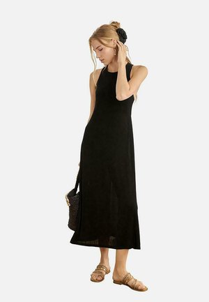 Mujer con vestido midi negro sin mangas, sandalias marrones, sosteniendo un bolso tejido, con el cabello rubio recogido y un accesorio negro para el cabello.