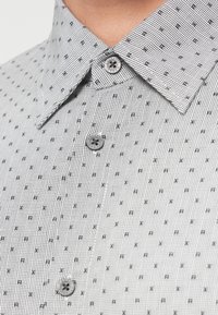 Κοντινό πλάνο λευκού πουκαμίσου με μαύρο καρό σχέδιο, μικρά γράμματα "R" και "X", και μάρκα Armani Exchange κουμπιά που κλείνουν τον γιακά και το μπροστινό μέρος.