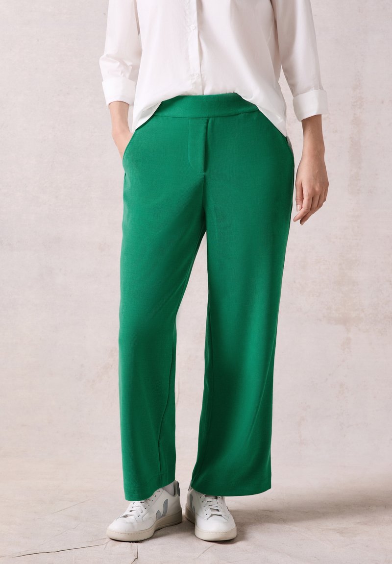 Gröna vida byxor med en slät textur, elastisk midja och sidofickor; kombinerat med vita sneakers och en vit skjorta med knappar.