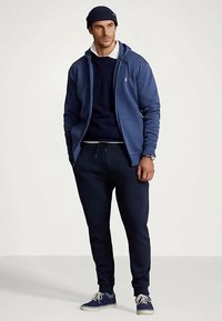 Άνδρας που φοράει μπλε σκουφί, μπλε φούτερ με φερμουάρ, ναυτικό πουλόβερ πάνω από λευκό πόλο, ναυτικές φόρμες, και μπλε αθλητικά παπούτσια, στέκεται με τα χέρια στις τσέπες.